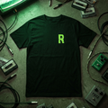 Black t-shirt with neon green glitch letter R chest print, flat lay on concrete with cassette tapes, guitar pedals, and a glowing CRT monitor